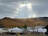 A serene landscape with a foreground of traditional yurts and modern buildings, set against a backdrop of a dense forest on a mountain slope. The sky is overcast with a partial opening that reveals sunlight breaking through.