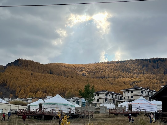 A serene landscape with a foreground of traditional yurts and modern buildings, set against a backdrop of a dense forest on a mountain slope. The sky is overcast with a partial opening that reveals sunlight breaking through.