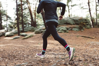 A person runs through a forested trail covered in brown leaves. The scene captures an autumn setting with tall trees and scattered boulders.