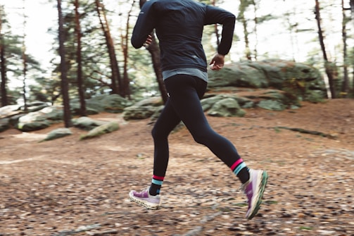 A person runs through a forested trail covered in brown leaves. The scene captures an autumn setting with tall trees and scattered boulders.