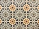 A symmetrical pattern of ceramic tiles featuring geometric shapes and floral motifs. The design consists of squares with intricate designs, combining circles, swirls, and stylized flowers in subdued colors. Each tile is neatly aligned to create a seamless repetition across the surface.