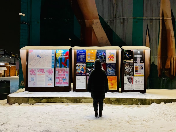 A person stands in front of illuminated outdoor boards displaying various posters with text and images in a snowy environment. The posters cover topics such as events and advertisements, and the scene is set against a background of a wall with mural art.