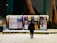 A person stands in front of illuminated outdoor boards displaying various posters with text and images in a snowy environment. The posters cover topics such as events and advertisements, and the scene is set against a background of a wall with mural art.