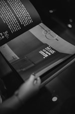 A black and white photograph of an open book held by a hand. The page on the left contains handwritten text, while the right page features printed text and a diagram with geometric shapes. The background is dark, highlighting the pages of the book.