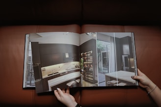 A person holding an open book displaying a modern interior design of a kitchen with dark cabinetry, a large refrigerator, contemporary lighting, and a small dining area. The page features a high-quality photograph with natural light coming through large windows.