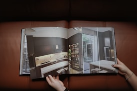 A person holding an open book displaying a modern interior design of a kitchen with dark cabinetry, a large refrigerator, contemporary lighting, and a small dining area. The page features a high-quality photograph with natural light coming through large windows.