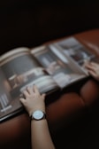 Two hands are seen flipping through the pages of a glossy magazine, with one of the arms wearing a minimalist watch. The blurred background suggests an indoor setting with soft, ambient lighting.