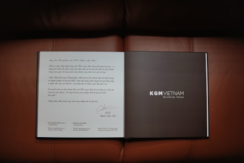 An open book or brochure is laid out on a brown leather surface. The left page contains text written in a decorative style, possibly a letter or introduction. The right page is dark with the text 'KGM VIETNAM' and 'Building Value' printed clearly.