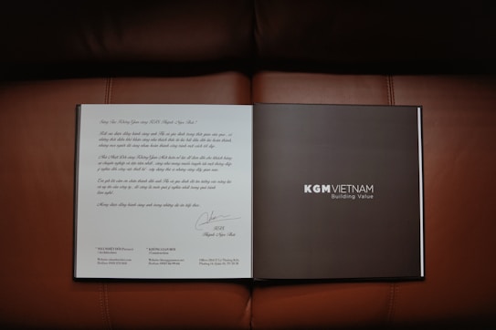 An open book or brochure is laid out on a brown leather surface. The left page contains text written in a decorative style, possibly a letter or introduction. The right page is dark with the text 'KGM VIETNAM' and 'Building Value' printed clearly.