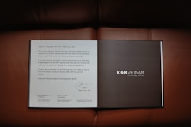 An open book or brochure is laid out on a brown leather surface. The left page contains text written in a decorative style, possibly a letter or introduction. The right page is dark with the text 'KGM VIETNAM' and 'Building Value' printed clearly.