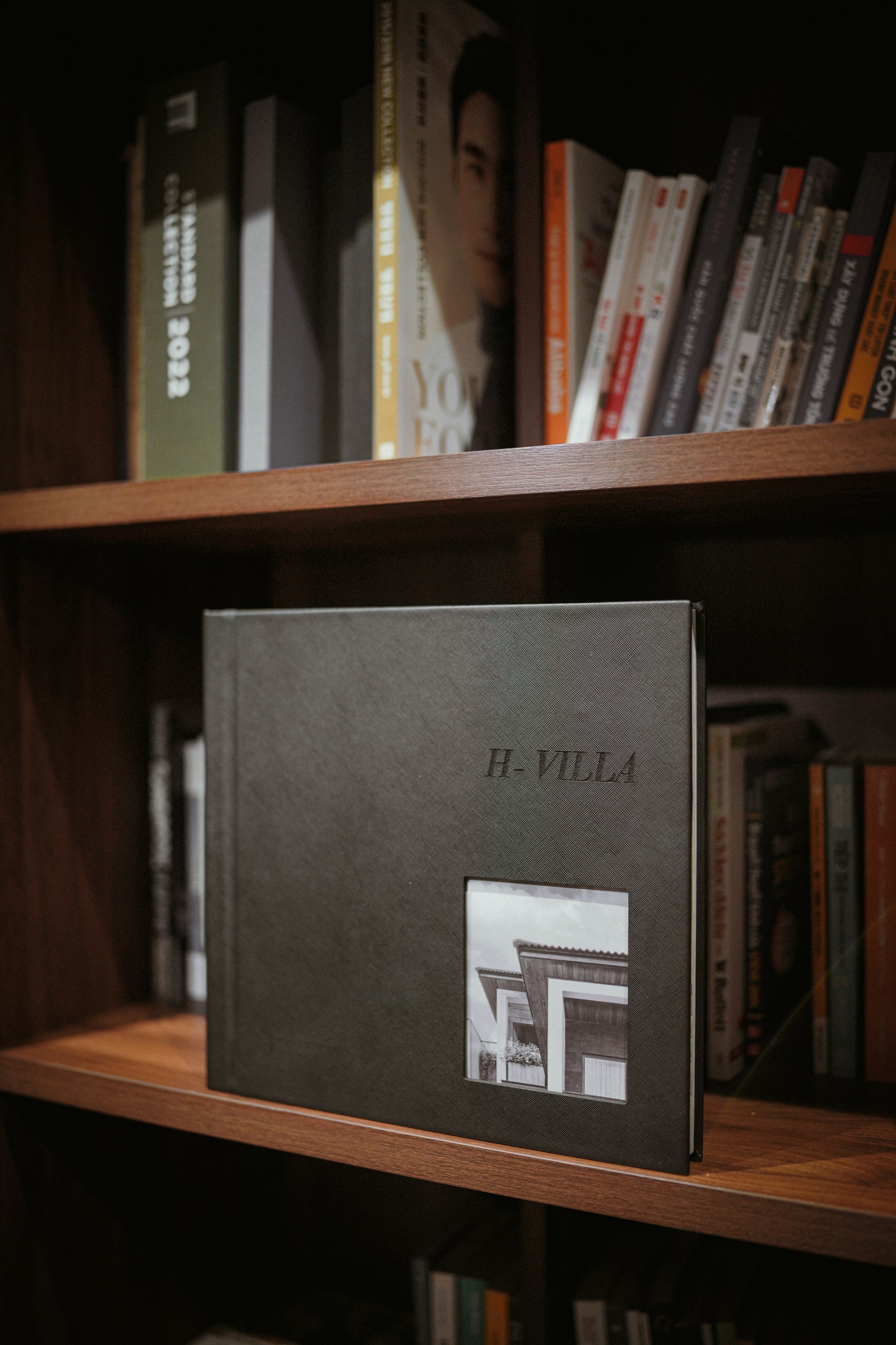 A bookshelf filled with various books, with a prominent photo album titled 'H-Villa' displayed in the center. The album is dark-colored with a small photograph of a building on its cover. The surrounding books are neatly arranged, with some visible titles.
