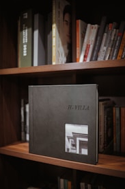 A bookshelf filled with various books, with a prominent photo album titled 'H-Villa' displayed in the center. The album is dark-colored with a small photograph of a building on its cover. The surrounding books are neatly arranged, with some visible titles.