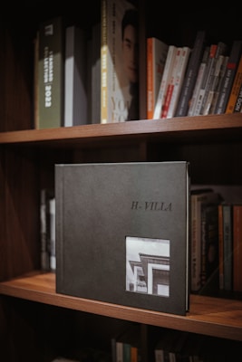 A bookshelf filled with various books, with a prominent photo album titled 'H-Villa' displayed in the center. The album is dark-colored with a small photograph of a building on its cover. The surrounding books are neatly arranged, with some visible titles.