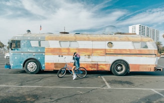 A vintage bus painted to resemble the American flag with a combination of stars and stripes is parked in an open area. A person dressed in a stylish outfit stands beside the bus, posing with a bicycle. The scene is set against a partly cloudy sky with a few palm trees and modern buildings visible in the background.
