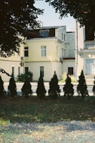 A row of neatly trimmed evergreen shrubs is lined up in front of a bright yellow two-story building with multiple windows and air conditioning units. The building is partially shaded by overhanging tree branches with lush green foliage. The foreground consists of a grassy area with scattered fallen leaves, indicating a serene urban environment.