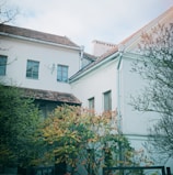 A residential building with white walls and a brown tiled roof is partially obscured by lush trees and shrubs. The windows have a traditional design, and the roof features a satellite dish. The foliage shows signs of autumn, with leaves turning shades of red and orange.