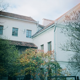 A residential building with white walls and a brown tiled roof is partially obscured by lush trees and shrubs. The windows have a traditional design, and the roof features a satellite dish. The foliage shows signs of autumn, with leaves turning shades of red and orange.