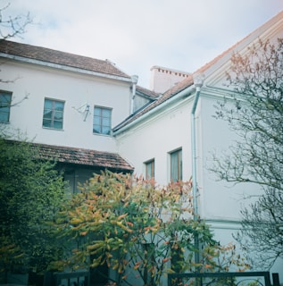 A residential building with white walls and a brown tiled roof is partially obscured by lush trees and shrubs. The windows have a traditional design, and the roof features a satellite dish. The foliage shows signs of autumn, with leaves turning shades of red and orange.
