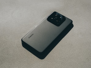 A modern smartphone with a sleek design lying flat on a textured surface. The device features a prominent camera module at the back and a logo indicating the brand. The lighting creates subtle shadows, adding depth and highlighting the phone's contours.