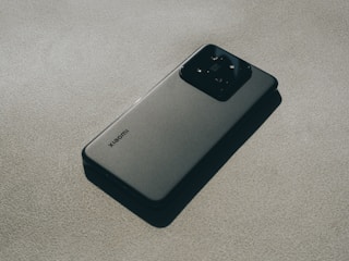 A modern smartphone with a sleek design lying flat on a textured surface. The device features a prominent camera module at the back and a logo indicating the brand. The lighting creates subtle shadows, adding depth and highlighting the phone's contours.