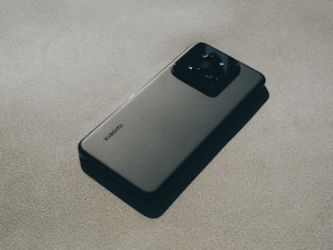 A modern smartphone with a sleek design lying flat on a textured surface. The device features a prominent camera module at the back and a logo indicating the brand. The lighting creates subtle shadows, adding depth and highlighting the phone's contours.
