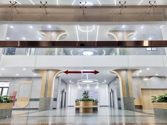 A modern interior space featuring circular architectural elements and a symmetrical design. Vertical columns with wooden accents merge into ceiling structures with embedded circular lights. Glass walls separate the different sections, and the floor is polished with a combination of light and dark shades. Green plants are strategically placed to add a touch of nature to the space.