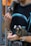 A man is holding two small monkeys, each with distinct white tufts on their ears. The first monkey is perched on his left hand, nibbling on food, while the second monkey rests on his arm, also holding food. The man is wearing a dark graphic t-shirt and a watch on his wrist. The setting appears indoor with a blurred background.
