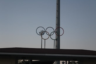 Five interlocking rings in different colors are mounted on a tall structure against a clear sky. A large, vertical pole and a partially visible stadium roof are in the background.