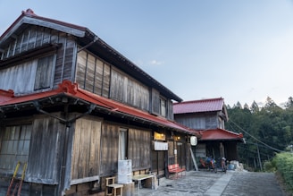 A traditional wooden Japanese house with red roof tiles, featuring rustic wooden walls and a serene outdoor setting. Bicycles are parked by the entrance, and there's a small red bench. The surrounding area is calm, with a backdrop of tall trees and a clear blue sky.