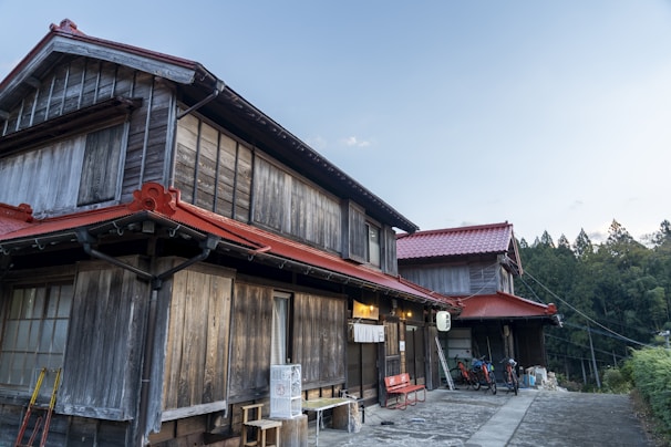 A traditional wooden Japanese house with red roof tiles, featuring rustic wooden walls and a serene outdoor setting. Bicycles are parked by the entrance, and there's a small red bench. The surrounding area is calm, with a backdrop of tall trees and a clear blue sky.