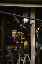 A person wearing a safety harness and helmet is standing on a ladder. They are working on a building, possibly installing or cleaning windows. The individual is equipped with various tools clipped to their belt, and there is a rope for additional safety.