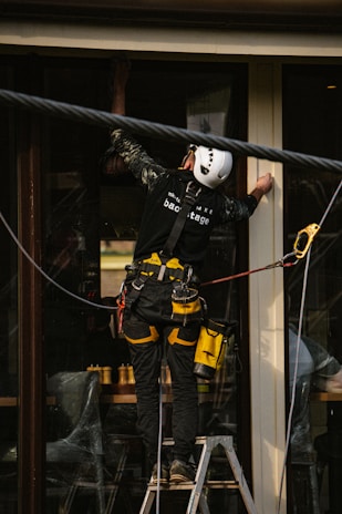 A person wearing a safety harness and helmet is standing on a ladder. They are working on a building, possibly installing or cleaning windows. The individual is equipped with various tools clipped to their belt, and there is a rope for additional safety.