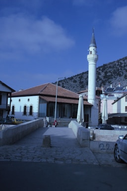A traditional Islamic mosque with a tall minaret stands prominently against a backdrop of blue sky and scattered clouds. Surrounding the mosque are several modern buildings and a mountainous landscape. A stone walkway leads to the mosque, where a few people are casually walking.