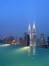 A city skyline at dusk with the Petronas Twin Towers prominently lit up against a clear blue sky. The foreground features an infinity pool, its surface reflecting the bright lights of the skyscrapers.