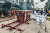 A traditional cultural ceremony is taking place on a road with a colorful, decorative cart at the center. Many people dressed in elaborate traditional attire are participating. Some are kneeling on the ground in respect or prayer, while others stand around observing. The scene is set outdoors surrounded by trees and rural buildings, creating a solemn and respectful atmosphere.