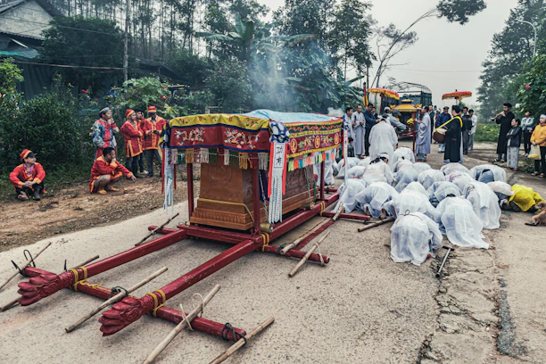 A traditional cultural ceremony is taking place on a road with a colorful, decorative cart at the center. Many people dressed in elaborate traditional attire are participating. Some are kneeling on the ground in respect or prayer, while others stand around observing. The scene is set outdoors surrounded by trees and rural buildings, creating a solemn and respectful atmosphere.