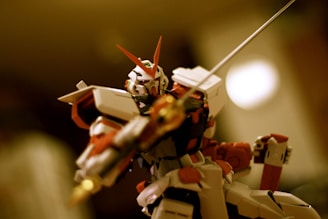 A detailed action figure resembling a robot or mecha, featuring a predominantly red, white, and black color scheme. The figure holds a weapon, possibly a sword or staff, emphasizing a dynamic and dramatic pose against a blurred background.