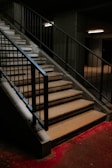 A concrete staircase with a metal railing is illuminated by overhead fluorescent lighting. The steps appear worn, leading up to a dimly lit area. The ground at the base is covered with a vivid red carpet or substance, creating a striking contrast with the muted tones of the stairs.
