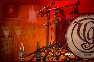 A drum set features prominently, highlighted by warm, ambient lighting. In the foreground, there are two beverage bottles, one clear and another dark green, placed on a surface. The background shows decorative elements like wall banners and posters, providing a musical and festive atmosphere. The lighting casts a soft glow, enhancing the overall warm tones.