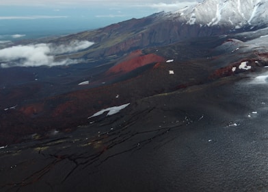 Aerial view of a volcanic landscape with dark, rugged lava fields stretching across the terrain. Snow-capped mountains are visible in the background, partially covered with patches of snow. A red volcanic cone stands prominently amidst the dark surface, suggesting recent volcanic activity. Clouds drift low in the scene, adding a misty atmosphere.