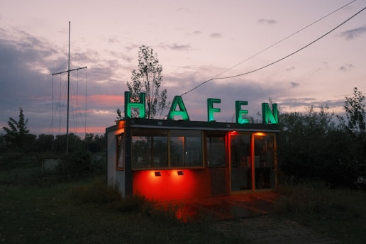 A small building with a sign on top displaying the word 'HAFEN' in bright green letters. The structure is illuminated by red lights at the entrance, set against a backdrop of overcast sky transitioning into dusk. The surrounding area is grassy with some trees and plants visible.