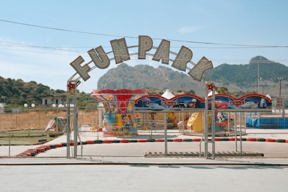 An entrance gate to a fun park with a large sign overhead. The area is fenced off, featuring various amusement rides with colorful decorations. Hills and trees are visible in the background under a clear blue sky.