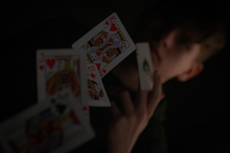 A person holding a playing card in their hand, with other playing cards floating in mid-air. The background is dark, highlighting the contrast between the cards and the surroundings.