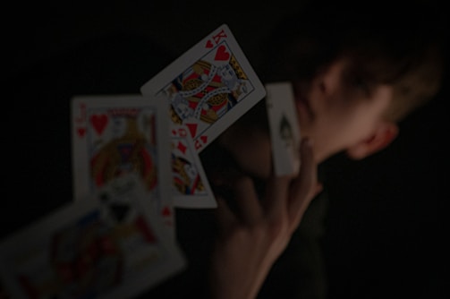 A person holding a playing card in their hand, with other playing cards floating in mid-air. The background is dark, highlighting the contrast between the cards and the surroundings.