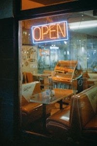 A dimly lit vintage diner is visible through the window, featuring brown leather booths and a neon 'OPEN' sign glowing in blue and orange. The atmosphere suggests a retro theme, with a jukebox in the background completing the nostalgic setting.