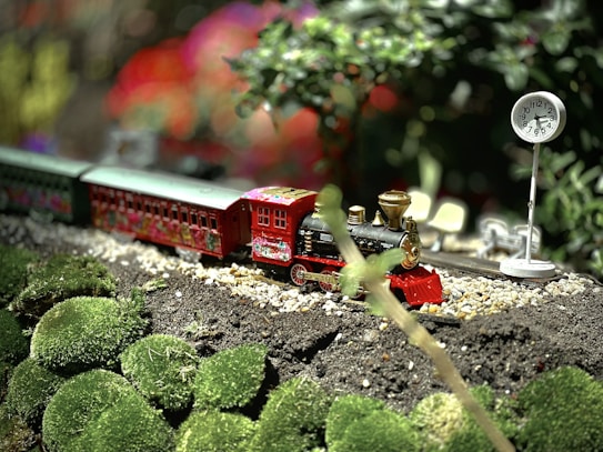 A miniature train runs through a scenic model landscape. It features a bright red engine and carriages with floral designs, set on a track surrounded by lush green moss and small white pebbles. A small clock on a pole stands nearby, indicating it is a train station setup. The background shows blurred, colorful lights or decorations, creating a vibrant atmosphere.