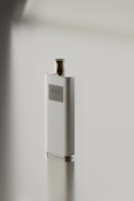 A sleek, rectangular perfume bottle with a metallic cap stands on a reflective surface. The label on the bottle reads 'DUA'. The minimalist design emphasizes simplicity and elegance.