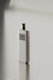 A sleek, rectangular perfume bottle with a metallic cap stands on a reflective surface. The label on the bottle reads 'DUA'. The minimalist design emphasizes simplicity and elegance.