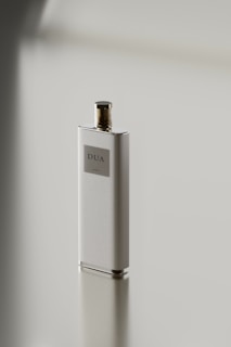 A sleek, rectangular perfume bottle with a metallic cap stands on a reflective surface. The label on the bottle reads 'DUA'. The minimalist design emphasizes simplicity and elegance.