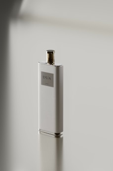 A sleek, rectangular perfume bottle with a metallic cap stands on a reflective surface. The label on the bottle reads 'DUA'. The minimalist design emphasizes simplicity and elegance.